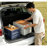 Person organizing an outdoor camping storage box in the trunk of a car, featuring wooden lids and snacks.